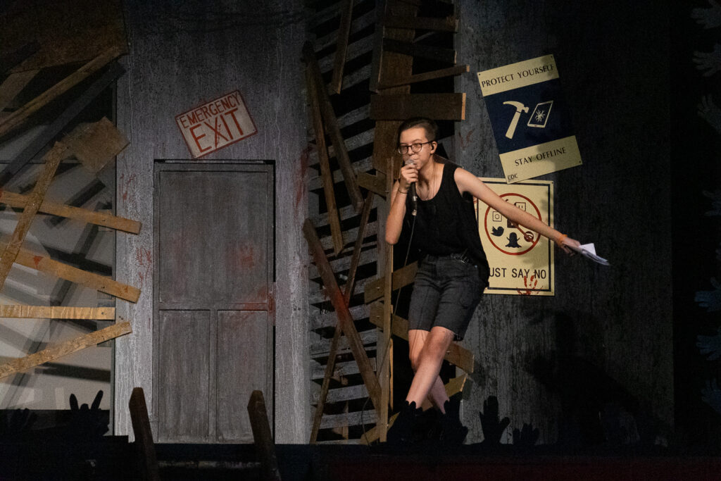 Performer on stage holding a microphone in front of a set designed with boarded-up walls, emergency exit sign, and warning posters.