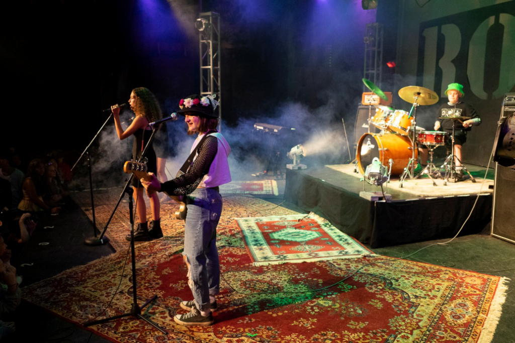 Picture5 – Long Lake Camp For The Arts Two female musicians sing into microphones while a guitarist plays on a colorful carpeted stage with smoke and a drum kit in the background.