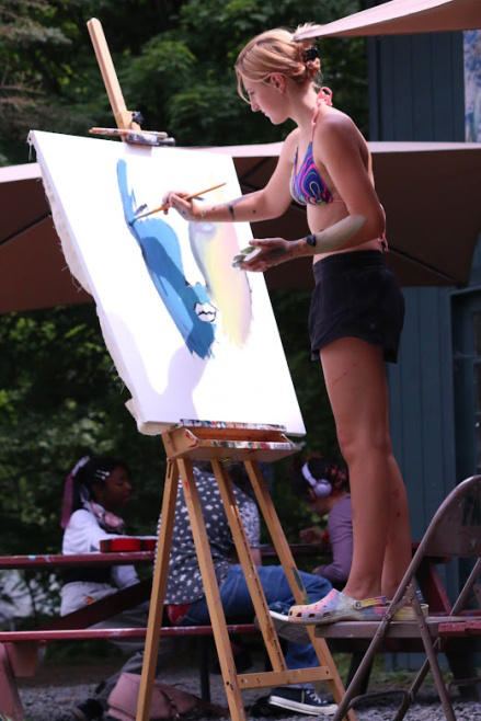 Picture24 – Long Lake Camp For The Arts A girl standing on a chair while painting a large canvas.