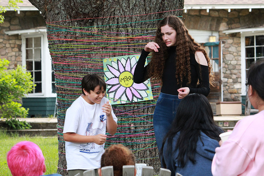 Picture1 – Long Lake Camp For The Arts Two campers perform a scene in front of fellow campers during a theater activity.